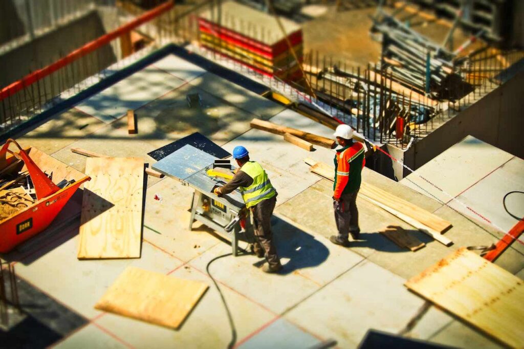 Two men wearing yellow and red PPE working on a construction site with cardboards and equipment scattered around